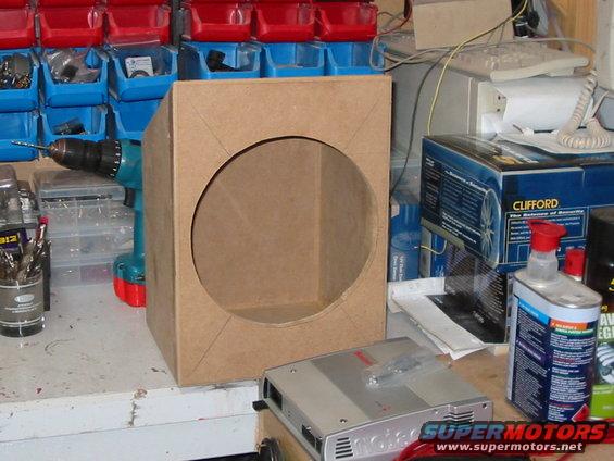 Cardboard speaker box with circular cutout, car amplifier, and drill on workbench