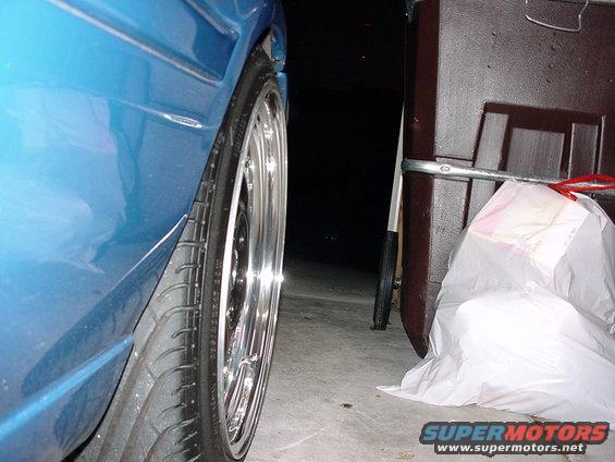 Blue car's rear wheel and fender, chrome rim, tire, next to a brown storage bin and white bag