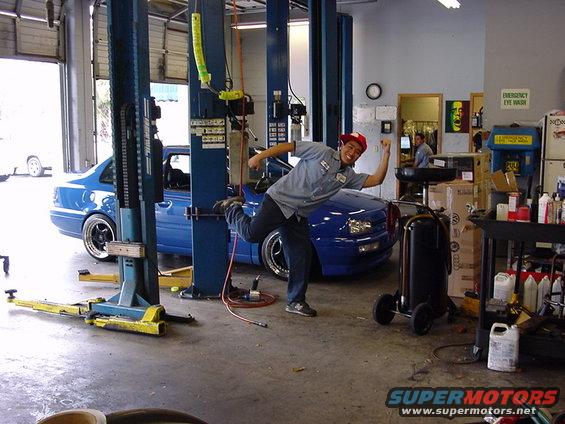 Man in red hat and work shirt posing with a blue 1998 Volkswagen Jetta in a garage with car lifts