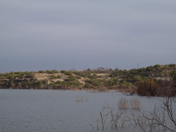 Oil pumpjacks on a distant hill overlooking a calm lake with bare branches in the foreground
