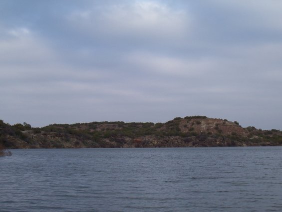 calm lake water in foreground with a tree-covered, rocky hill in the background under a cloudy sky