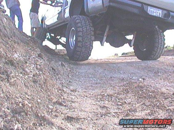 Off-road vehicle with large tires climbing a dirt mound, showing undercarriage and rear axle