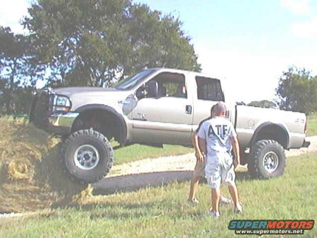 Lifted pickup truck with large tires on a grassy incline, two people standing nearby