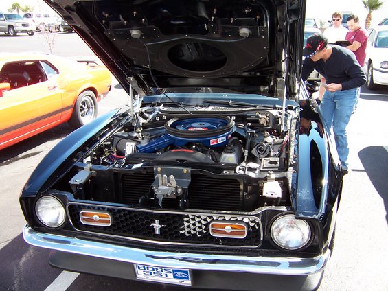 Black 1971 Ford Mustang Boss 351 front view, hood open, showing engine. Orange Mustang in background