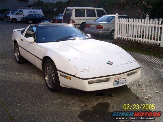 White Chevrolet Corvette parked outside with a black targa top. Other vehicles and a white fence are in the background