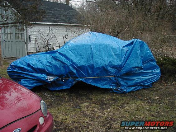 Red car hood in foreground, blue tarp covers another vehicle in yard with garage behind