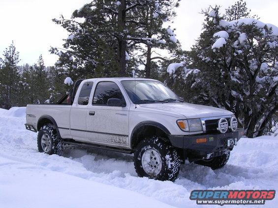 White 1999 Toyota Tacoma pickup truck driving in snow with aftermarket bumper and lights