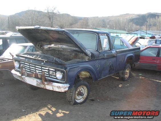 Old Ford F-series truck with hood open, parked in a junkyard. It has a flat tire and some graffiti on the side