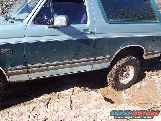 Side view of a light blue and dark green Ford Bronco stuck in mud