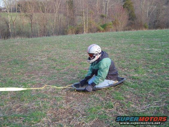 Person in sled with helmet and goggles, pulled by yellow rope across grassy field