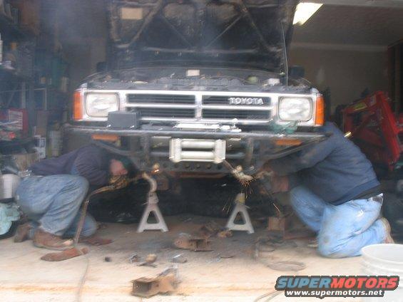 Toyota truck with hood open, front bumper being welded by two people. Sparks fly from welding