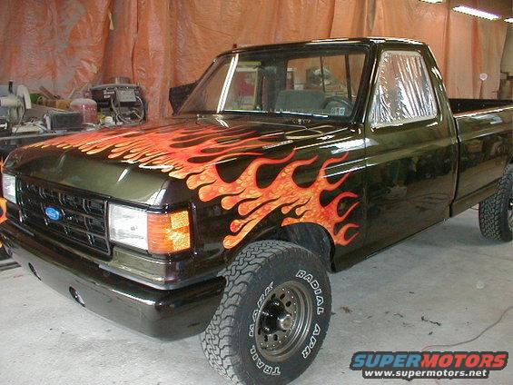Dark green Ford F-150 truck with orange and red flame decals on the hood and fender. The truck is in a garage with orange tarps in the background
