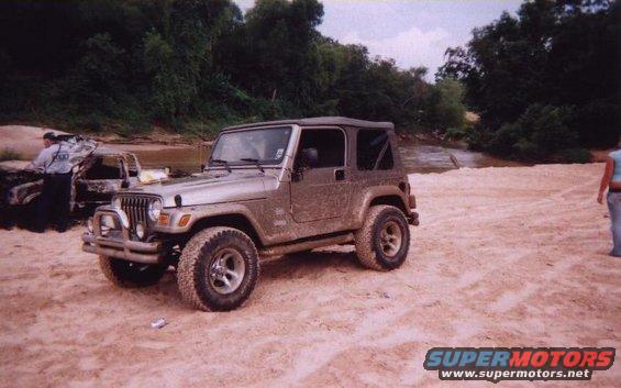 Silver Jeep Wrangler on sandy ground near a river, with a burnt vehicle in the background