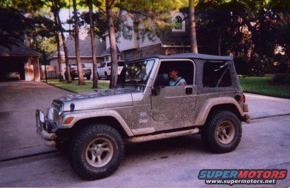 Silver Jeep Wrangler with a soft top, parked on a driveway. A person is visible in the driver's seat