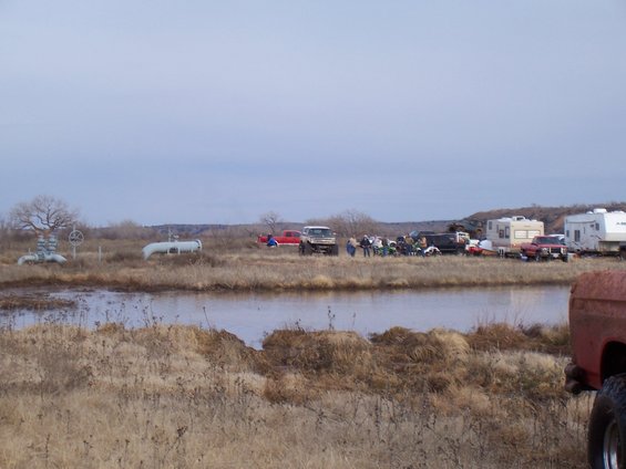 Several off-road trucks, campers, and people gathered near a muddy area with industrial pipes in the background