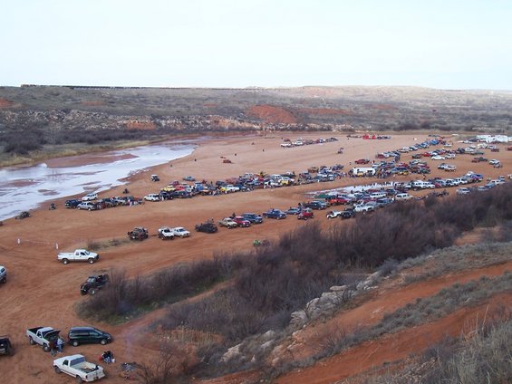 Many cars and trucks parked on a dirt area near a river, with a train visible on a distant ridge
