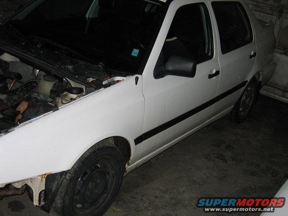 White sedan, hood open, showing engine components. Black trim along doors, black side mirror. Some rust on front fender