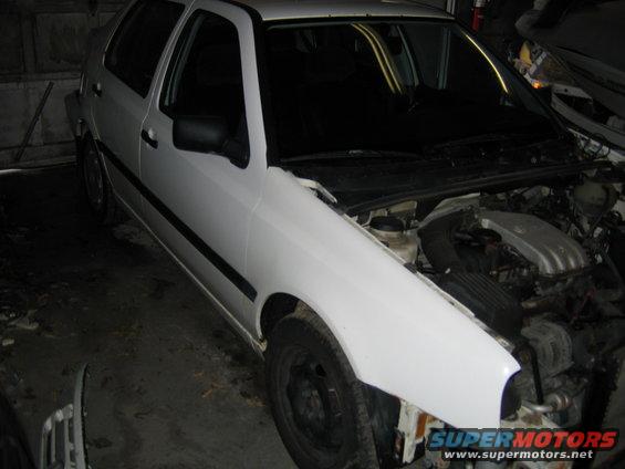 White hatchback with hood open, showing engine components. Front bumper is missing