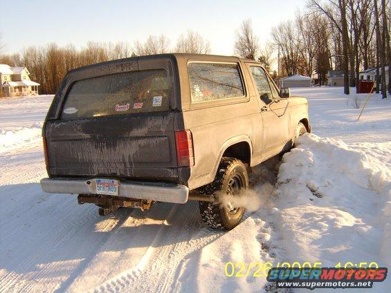 Rear view of a 1985 Ford Bronco stuck in snow, with exhaust smoke visible