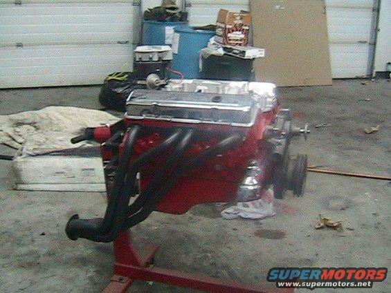 Red V8 engine with chrome valve covers and black headers on a stand