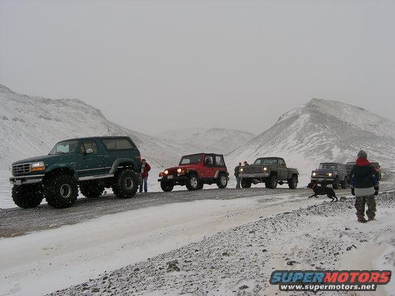 Several modified off-road vehicles, including a green Ford Bronco and a red Jeep, drive on a snow-covered road with mountains in the background