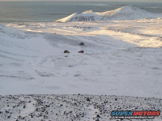 Three SUVs driving on a snow-covered hill with mountains and ocean in the background