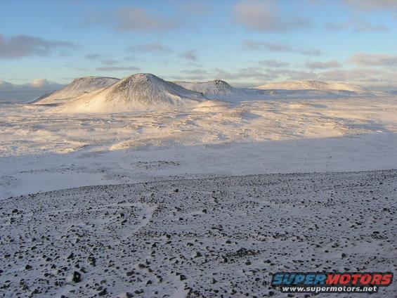 Snow-covered terrain with rolling hills and mountains under a blue sky with clouds