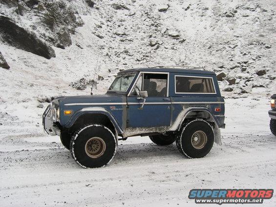 Blue Ford Bronco with big tires on snowy ground, another vehicle partially visible to the right