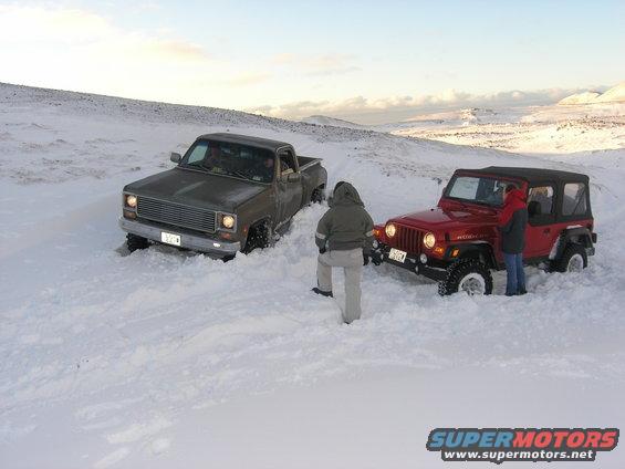 Red Jeep Wrangler and a dark pickup truck stuck in deep snow, with two people standing nearby