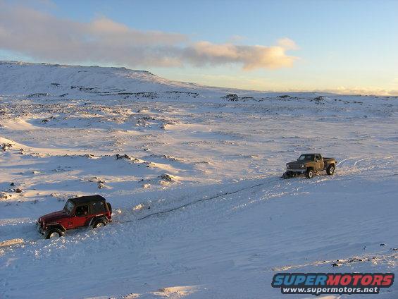 Red Jeep and brown pickup truck driving in snow, with a tow rope between them