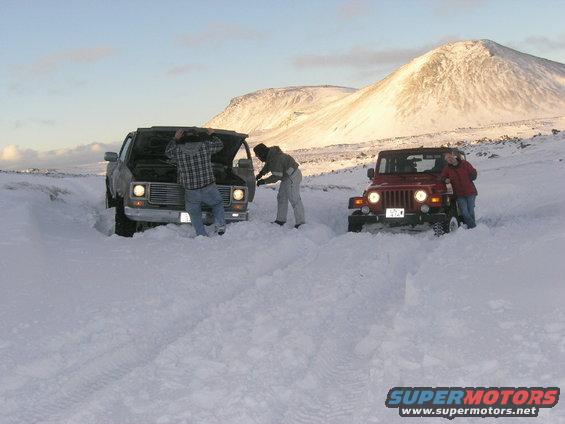 Two people working on a truck with its hood up, stuck in deep snow next to a red Jeep