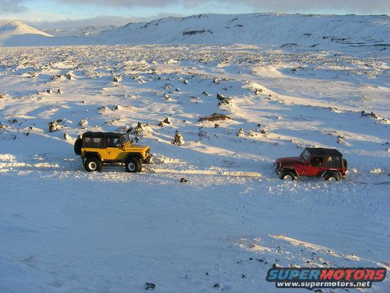 Yellow and red Jeeps driving in a vast, snow-covered, rocky terrain with mountains in the background
