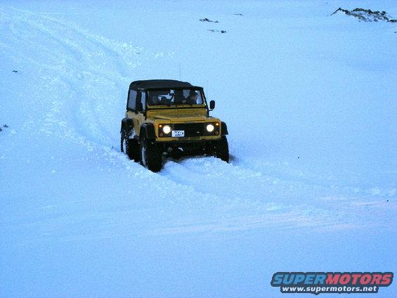 Yellow Land Rover Defender driving through snow, front view. License plate is visible