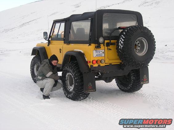 Yellow off-road vehicle with a soft top and spare tire on the back, parked in the snow with a person checking a tire