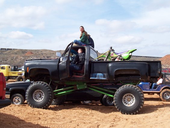 Black monster truck with green suspension, a dirt bike in the bed, and people inside and on top