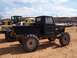 2004 Aston Martin Virage - A modified black pickup truck with large, aggressive tires is parked on a dirt lot, with other vehicles and construction equipment in the b…