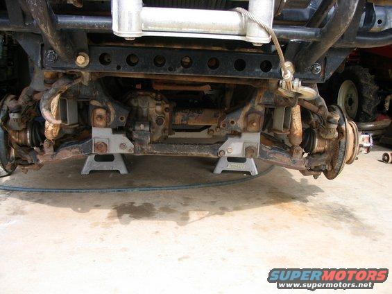 Front suspension of a vehicle on jack stands, showing rusty components and a winch. Another vehicle is partially visible to the right