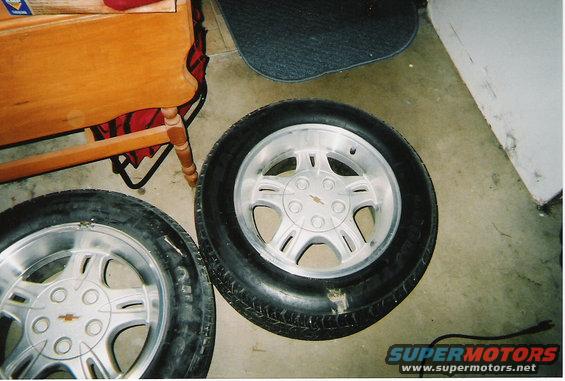 Two aluminum alloy rims with tires, one with a Chevrolet emblem, on a concrete floor next to wooden furniture