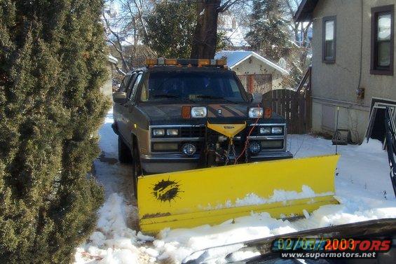 1990 Chevrolet Suburban with a yellow snow plow attached, front view, dark color, in snow