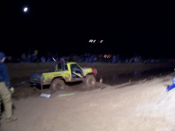 Yellow pickup truck with roll cage stuck in mud at night, with spectators in the background