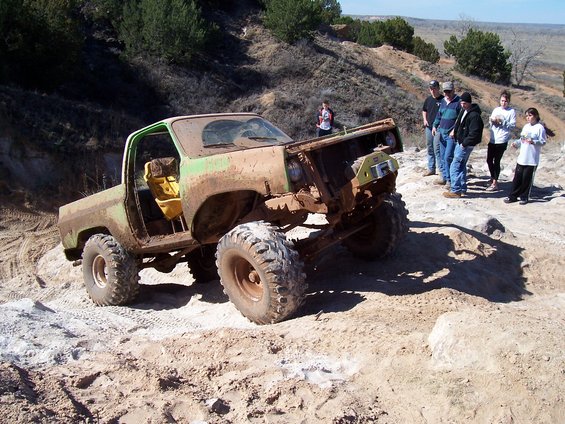 Muddy, lifted pickup truck with no hood, parked on rocky terrain. People stand nearby