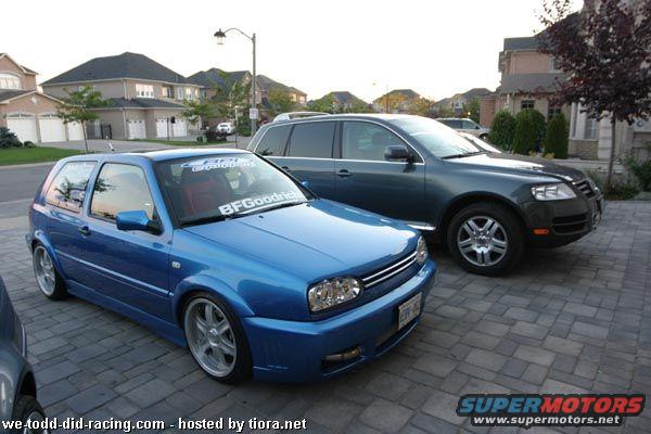 Blue VW Golf GTI parked next to a dark SUV. BFGoodrich sticker on windshield