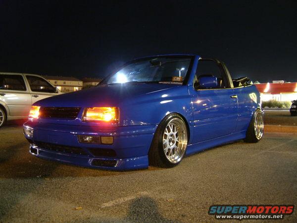 Blue convertible with aftermarket wheels parked at night. Another car is visible to the left