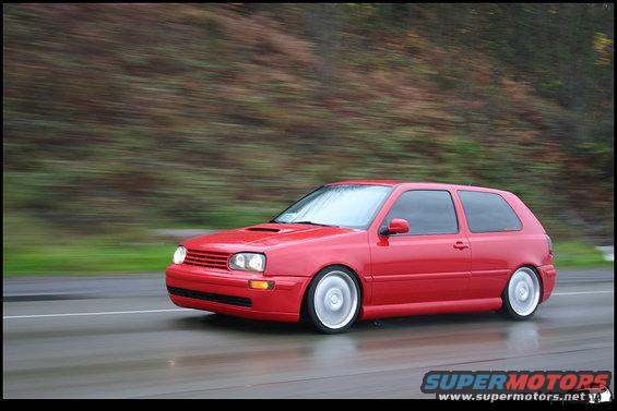 Red hatchback car with lowered suspension and aftermarket wheels driving on a wet road