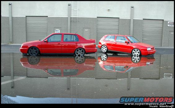 Two red Volkswagen Jettas, one sedan and one hatchback, parked side-by-side with their reflections in a wet lot