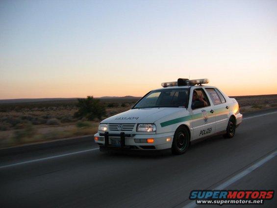 White police car with green stripe and light bar driving on highway at sunset