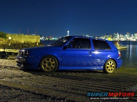 Blue hatchback with gold wheels parked by the water at night