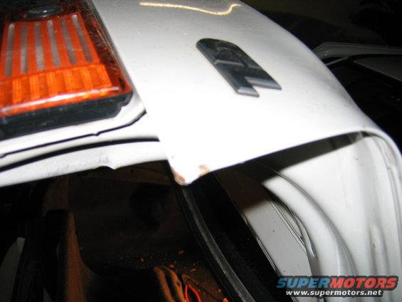 White car hood with a black emblem and orange turn signal. Small rust spot visible