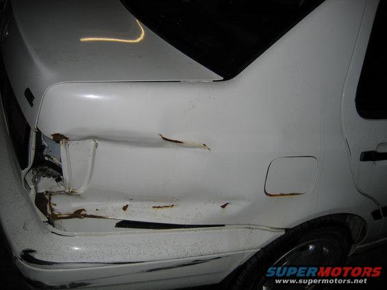 White sedan's rear passenger side has significant damage and rust near the taillight and along the body panel