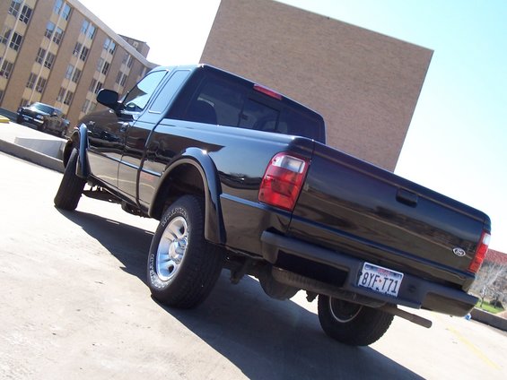 Black 2003 Ford Ranger pickup truck parked, rear view. Another car is visible in the background